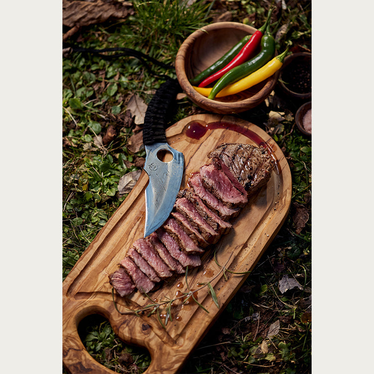 Almazan Kitchen artisanal olive wood cutting board with juicy sliced steak and Predator knife, surrounded by fresh peppers and rustic outdoor setting.