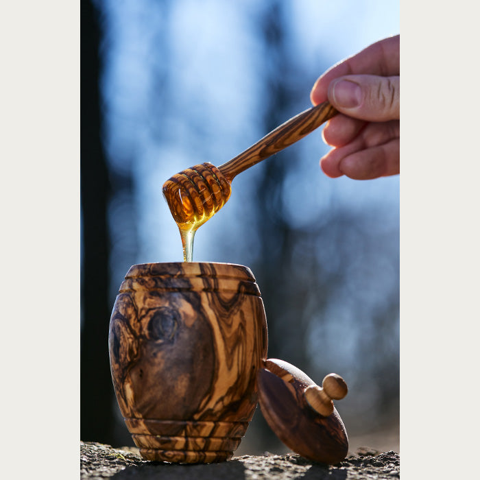 Golden honey being twirled on dipper over carved olive wood jar, with a blurred nature background.