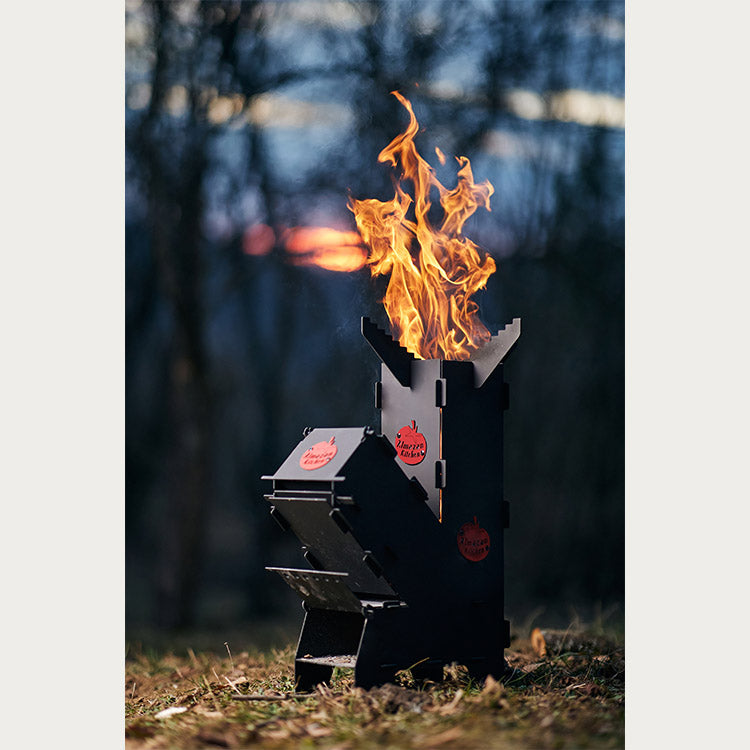 Foldable Almazan Kitchen Rocket Stove with vibrant flames burning, standing on grass with a blurred forest background during dusk.