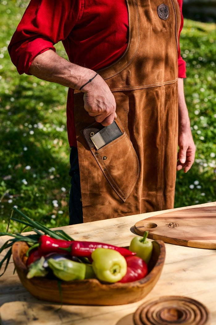 Man standing behind a wooden table with vegetables and a cutting board on it, putting a chef knife in a pocket of a leather apron.