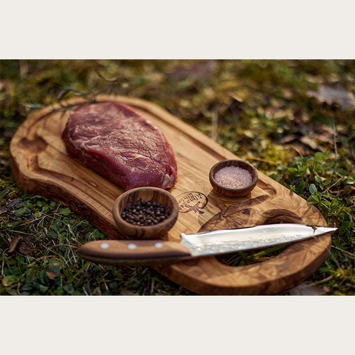 Almazan Kitchen's olive wood cutting board featuring a raw steak ready for seasoning, with Himalayan salt, whole black peppercorns, and a professional chef's knife on a outdoor backdrop.