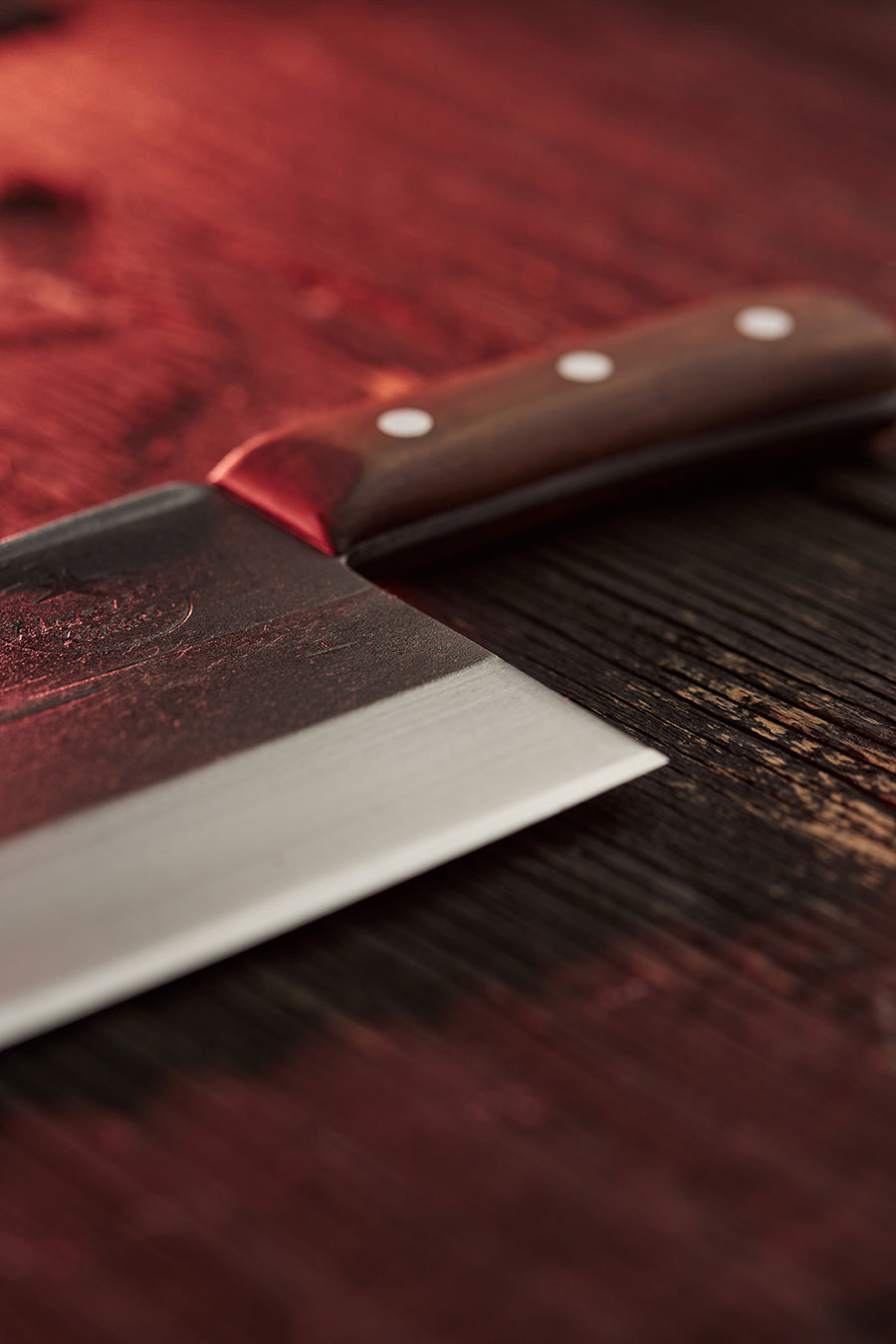 Blade and handle of the Serbian Chef Knife in focus, on a wooden table illuminated by a faint red light.