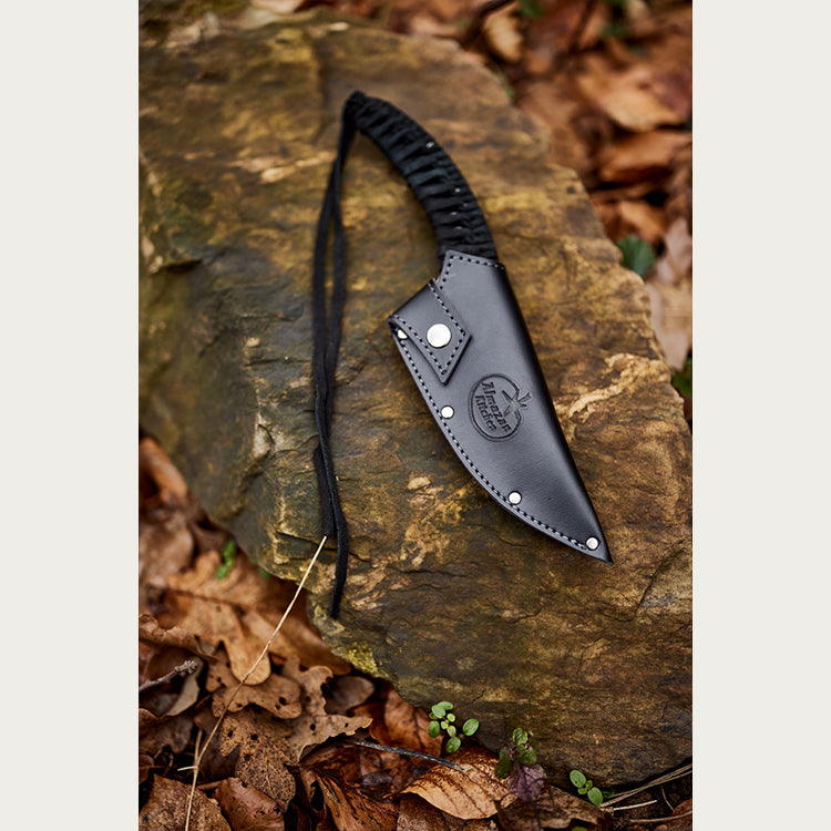 Almazan Kitchen Predator Knife in a black leather sheath set on a stone in a forest. 