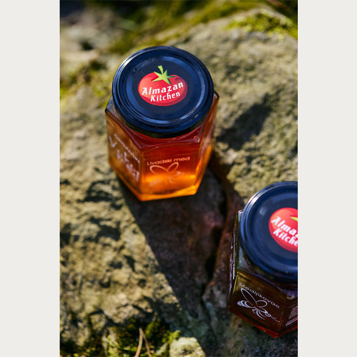 Two jars of honey with the Almazan Kitchen logo on the lid set on a stone with the sun shining on them.