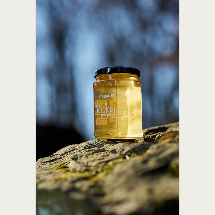 A jar of honey set on a stone, lit up by rays of sun with a nature backdrop.