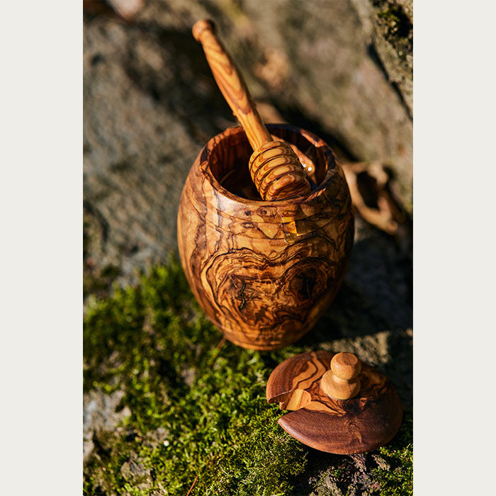 Artisanal olive wood honey jar with honey dipper, in an outdoor setting with the jar lid set down on moss.