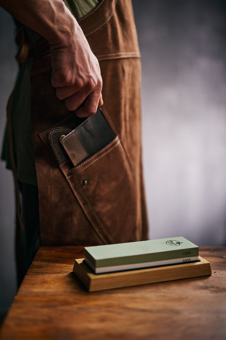 Sharpening stone set on a table and a man pulling out a chef knife from his leather apron pocket.