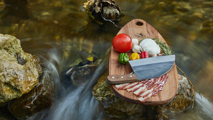 Almazan Kitchen cutting board with vegetables, meat and a Serbian Chef Knife on it, set on a stone in the middle of a creek.