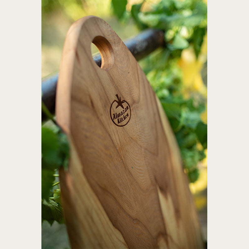 Walnut Cutting Board surrounded by leaves with the focus on the Almazan Kitchen logo