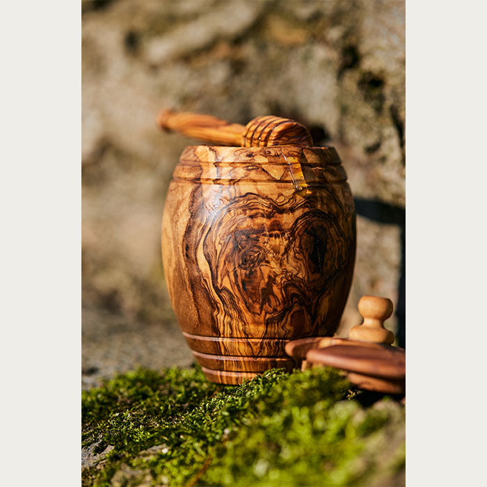 Artisanal olive wood honey jar with honey dipper set down on moss with the sun shining on it, with a lid in the foreground.