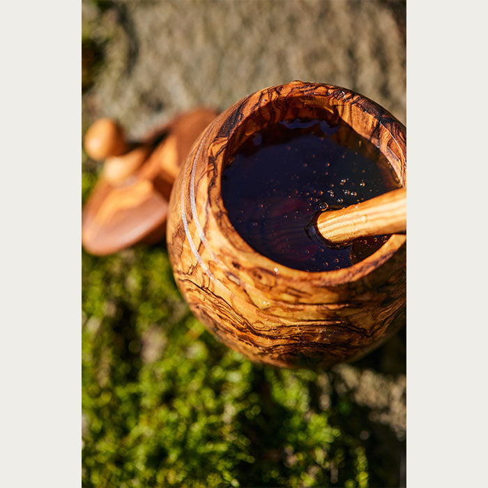 Wooden jar full of honey with a dipper inside of it, with a nature backdrop and the lid out of focus behind it.