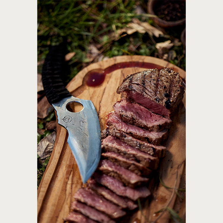 A cut up piece of steak on a cutting board next to a predator knife. 