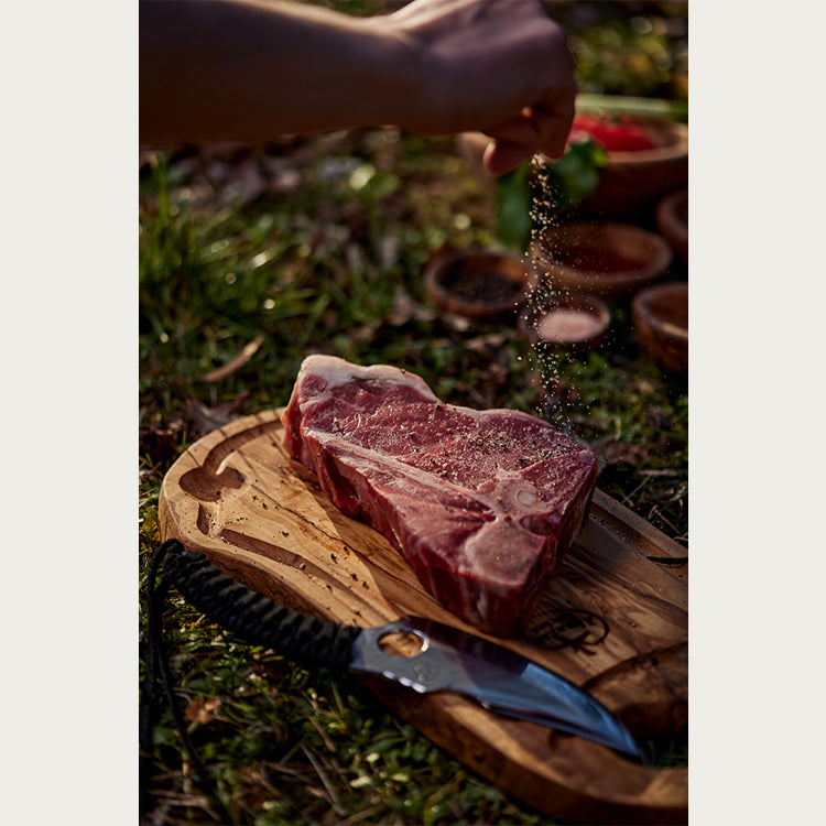 Person seasoning a piece of steak on a wooden cutting board, with a predator knife next to it. 