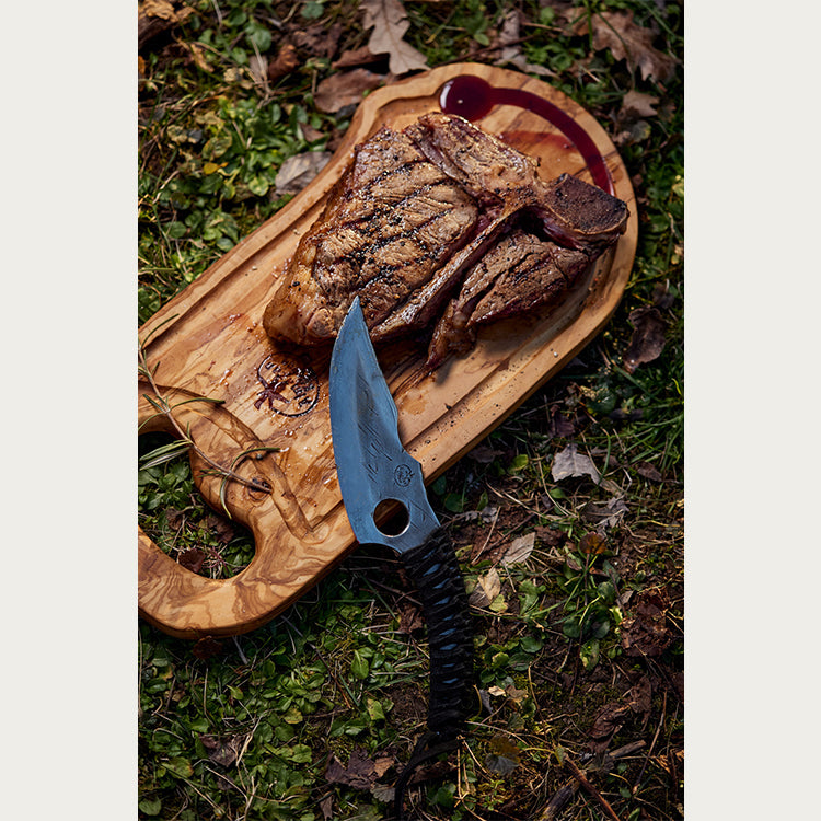 A cooked piece of steak on a cutting board with the tip of a predator knife set on top of it. 