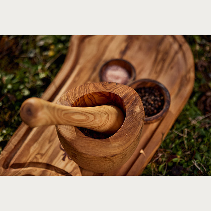 Overhead view of a handcrafted wooden mortar and pestle on a serving platter, with spices in two small bowls, set upon a grassy background in daylight.
