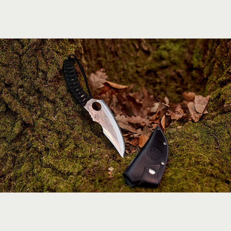Almazan Kitchen Predator Knife leaning on a mossy tree trunk with a black leather sheath in front of it. 
