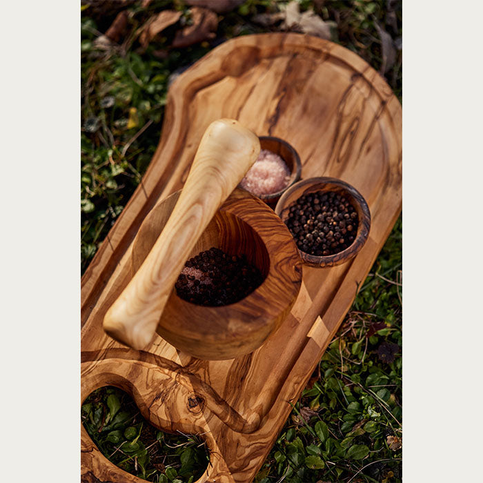 Overhead view of a wooden mortar and pestle on a cutting board, with spices in two small bowls, set upon a grassy background.