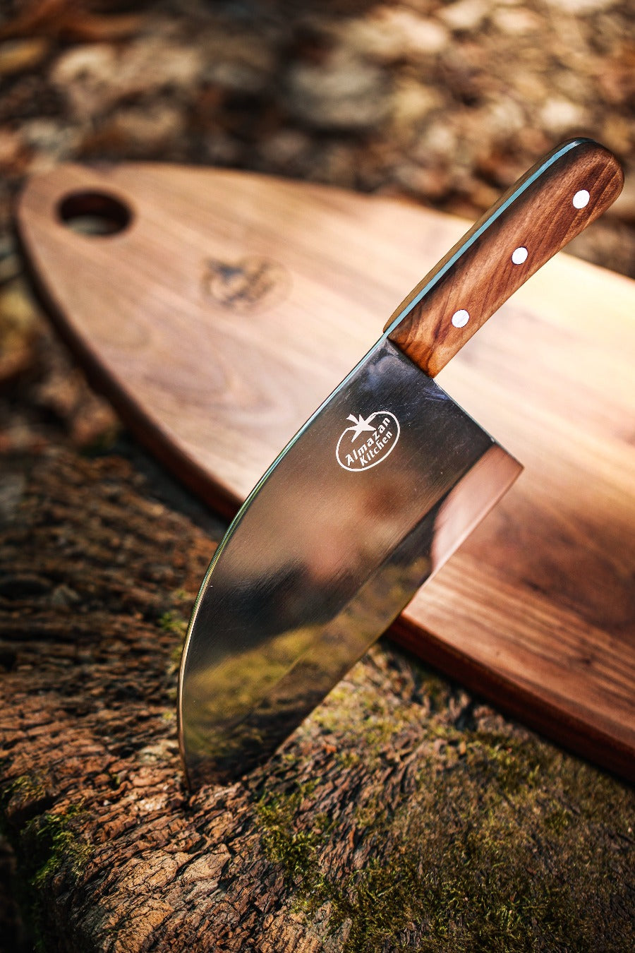 Almazan Kitchen Promaja Knife lodged into a tree trunk with a cutting board behind it.
