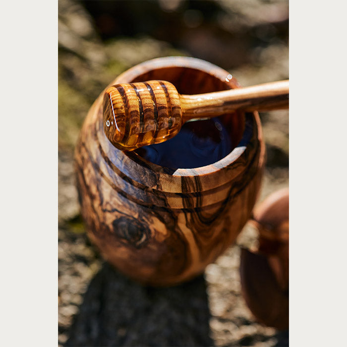 Artisanal olive wood honey jar with honey dipper drizzling honey, in an outdoor setting with the dipper in focus.