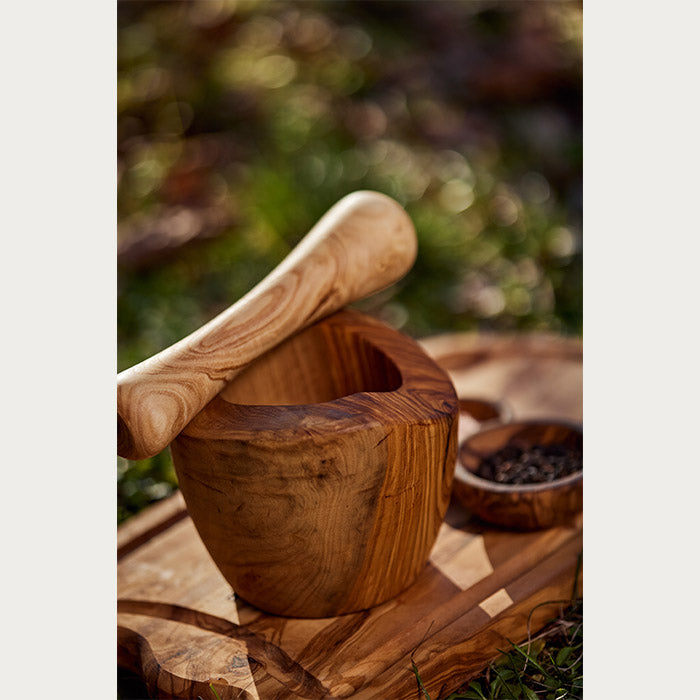 Artisan wooden mortar and pestle on a cutting board with spices in the background, set on a natural grass surface with soft-focus sunlight filtering through.