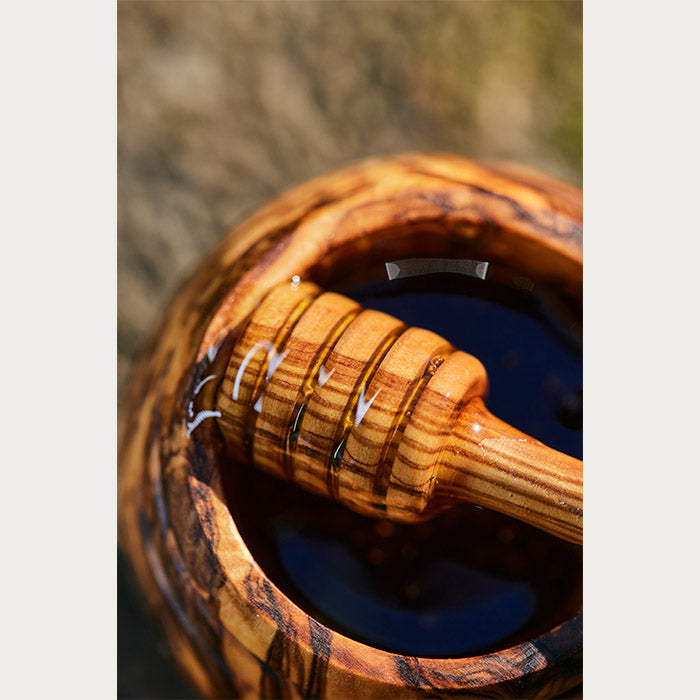 Honey dripping from a wooden dipper into a wooden jar with intricate grain patterns.