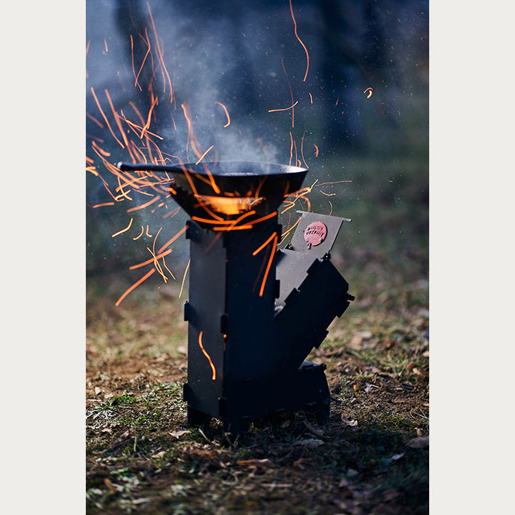 A pan on top of the Almazan Kitchen Rocket Stove with fire burning in it set in a forest.