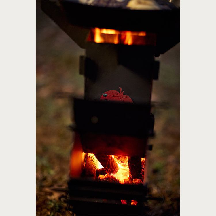 Almazan Kitchen Foldable Rocket Stove with a glowing firebox, against a soft, natural background in low light conditions.