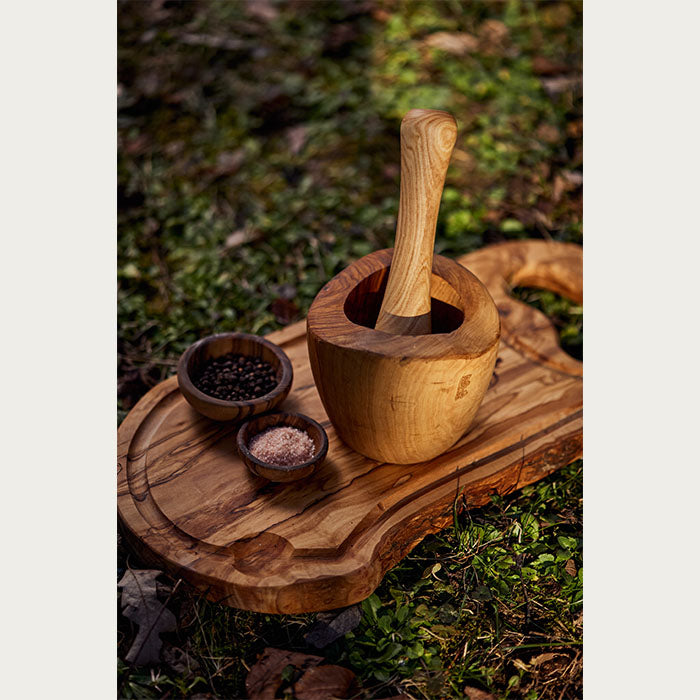 A wooden mortar and pestle set on a cutting board together with two small bowls full of spices, set upon a grassy background with rays of sun shining on them.