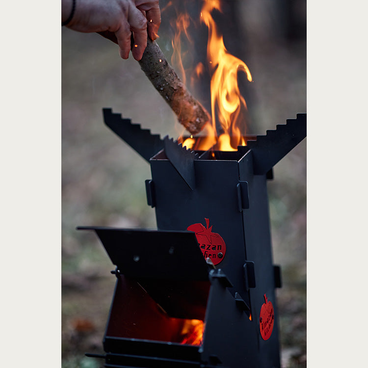 Man putting firewood into the Almazan Kitchen Rocket Stove with flames burning out of it.