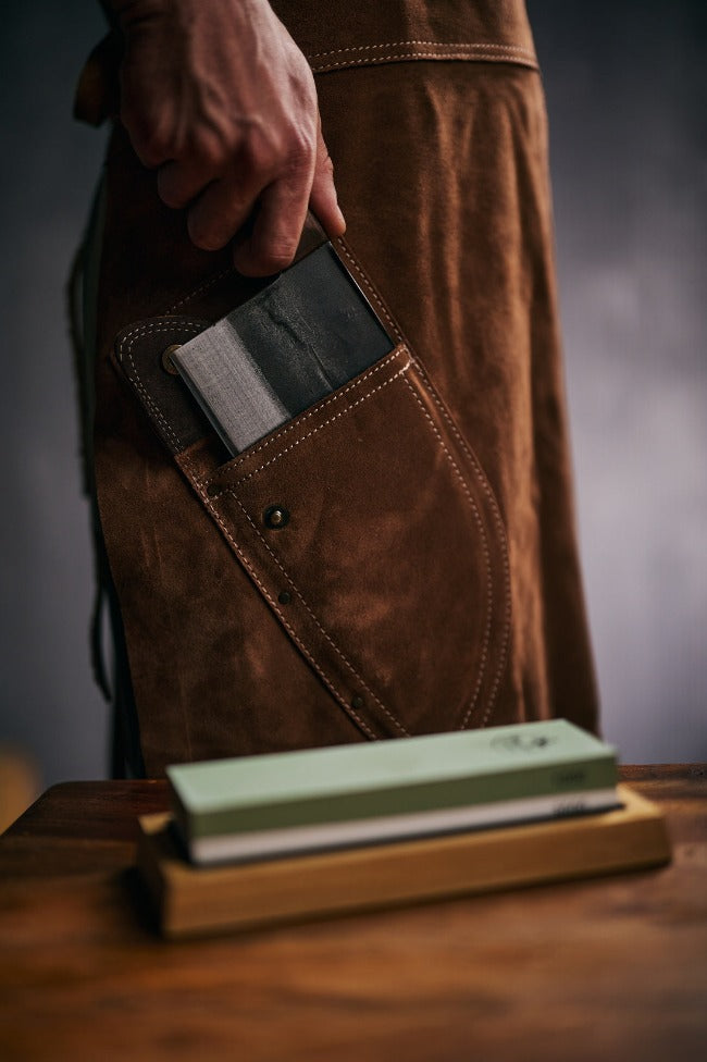 Sharpening stone set in the foreground with a person pulling out a chef knife from a pocket of a leather apron.